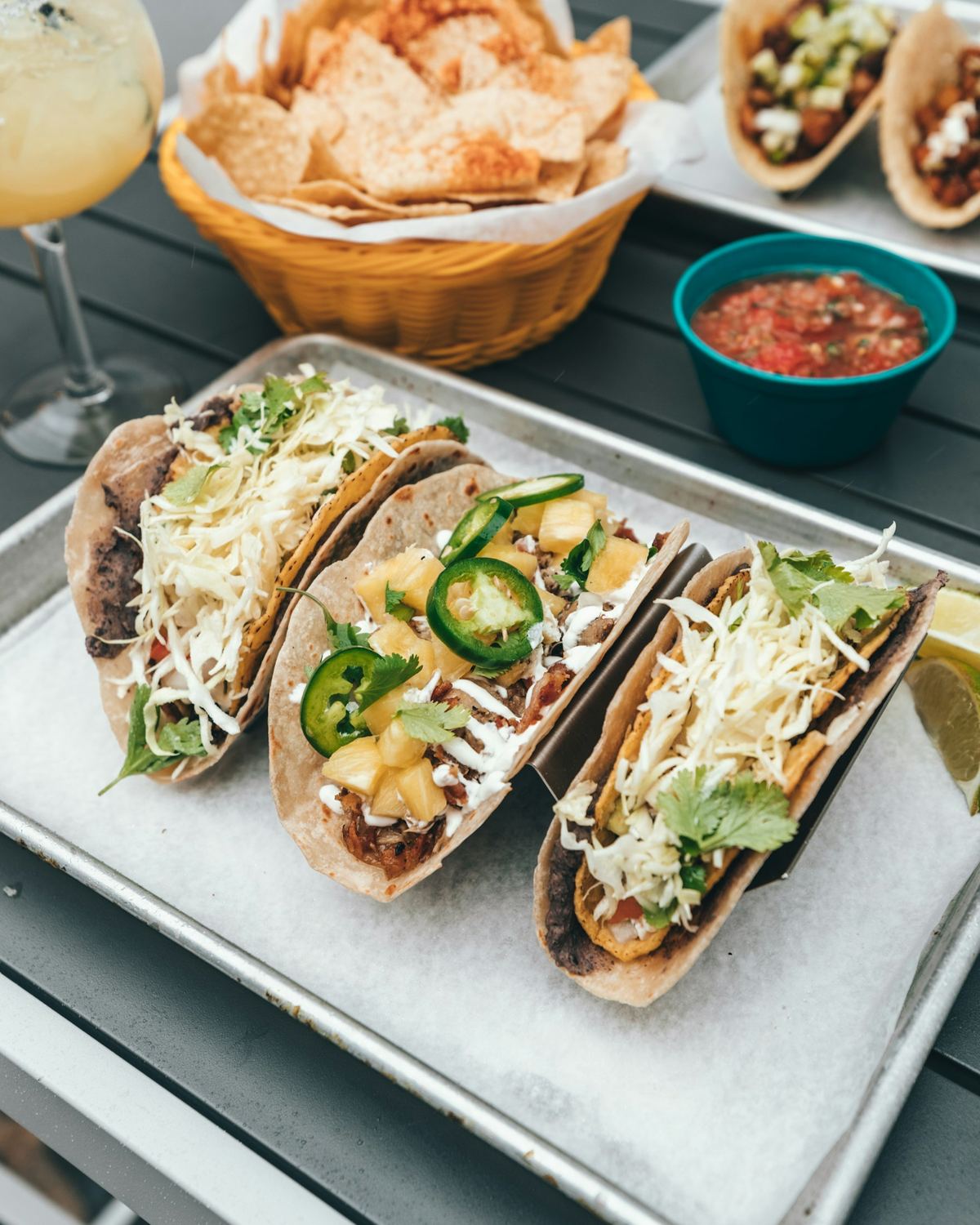 Three loaded tacos on a metal tray beside chips and a bowl of red salsa