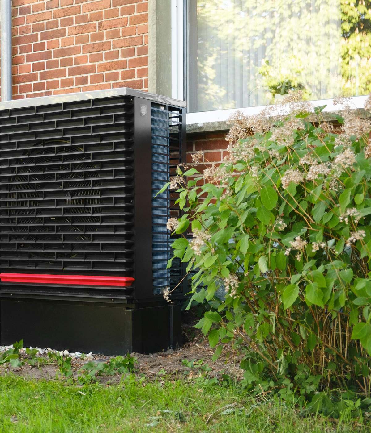A dark outdoor heat pump unit beside a brick wall and garden shrubs